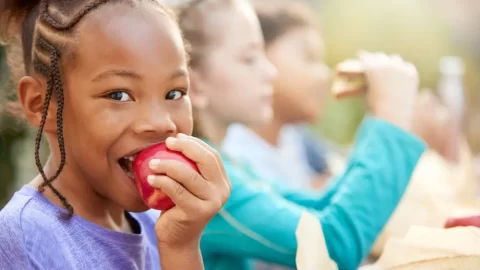 criança negra comendo maçã, com duas crianças a seu lado comendo